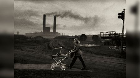 Black and white photograph of a woman pushing a pram along a bleak industrial road, with factory chimneys smoking in the background.