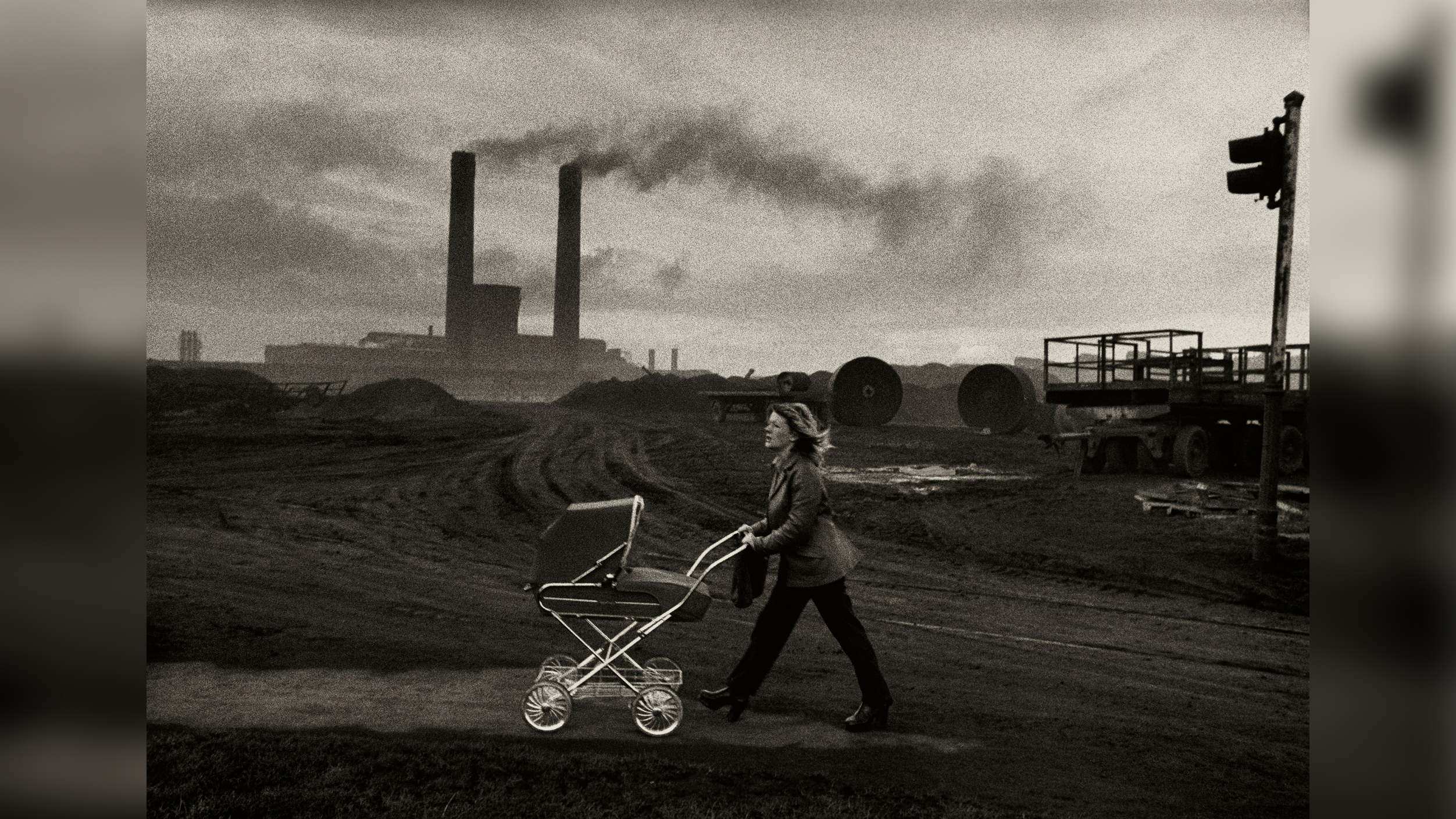 Black and white photograph of a woman pushing a pram along a bleak industrial road, with factory chimneys smoking in the background.