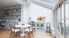A bright and tidy sunroom with wooden flooring, a white dining table, and brown wooden chairs. Behind is a cabinet with two matching lamps on top, and a white vase filled with orange flowers. To the left a white kitchen space is seen with neatly organized open shelving. To the right are bright windows.