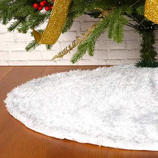 A white faux fur tree skirt under a faux Christmas tree, on a dark wooden floor against a white brick wall.