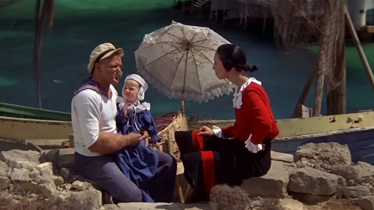 Robin Williams sitting with a baby on his knee, next to Shelly Duvall in Popeye.