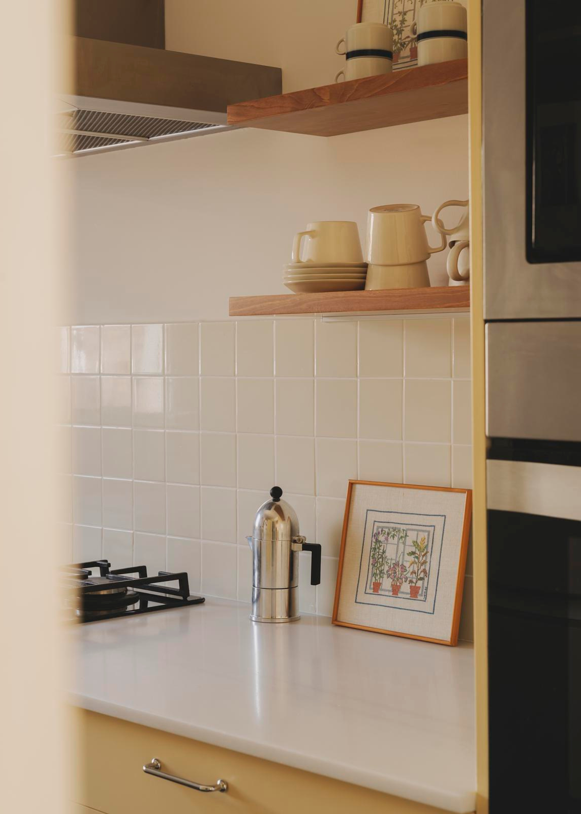 A close-up of a kitchen with a coffe maker, a framed print, and wooden shelving holding cups and saucers