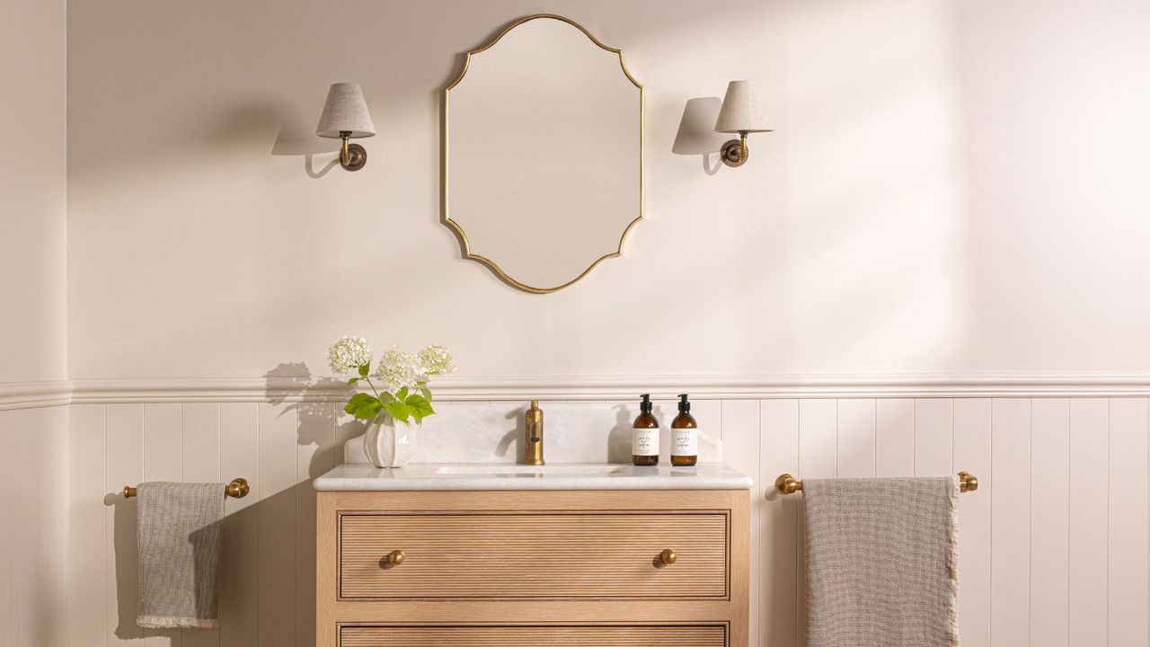 A wooden bathroom vanity until with drawers and a single sink. A gold rimmed mirror above with two wall sconces. Towels hanging on brass hardware either wide of the unit. 