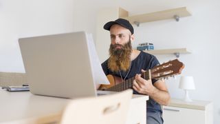 Bearded man plays acoustic guitar in front of his laptop
