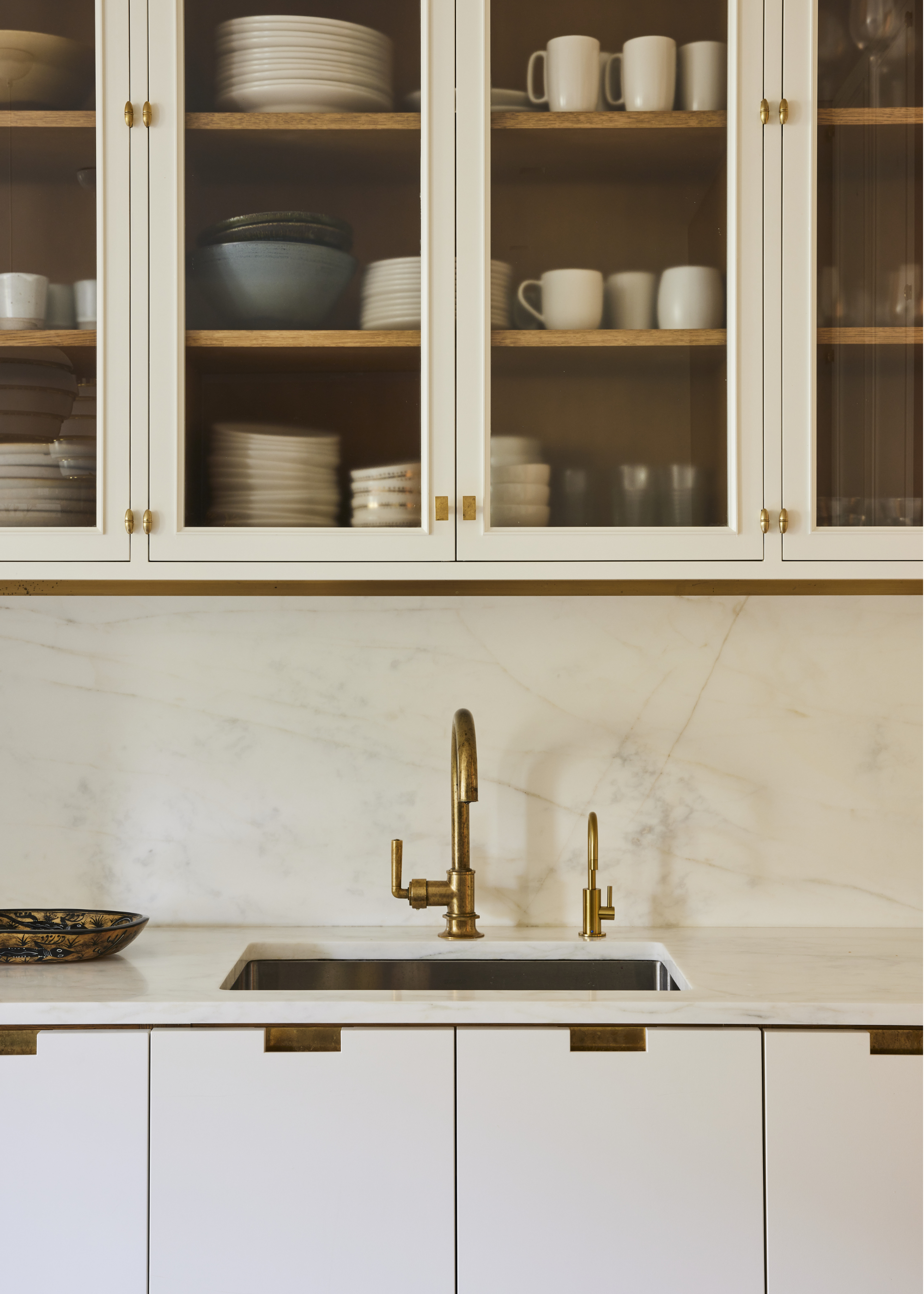 A cream kitchen with a sink and golden tap as well as glass fronted cabinets