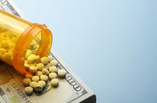 Pills spill out onto a one hundred dollar bill from an open prescription medication bottle. Ample room created by light blue background for copy and text.