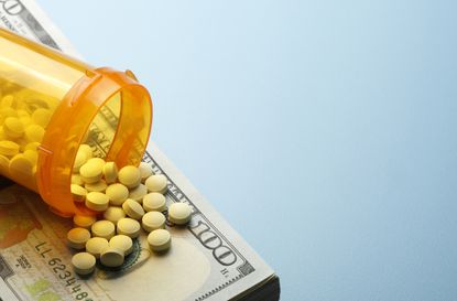 Pills spill out onto a one hundred dollar bill from an open prescription medication bottle. Ample room created by light blue background for copy and text.