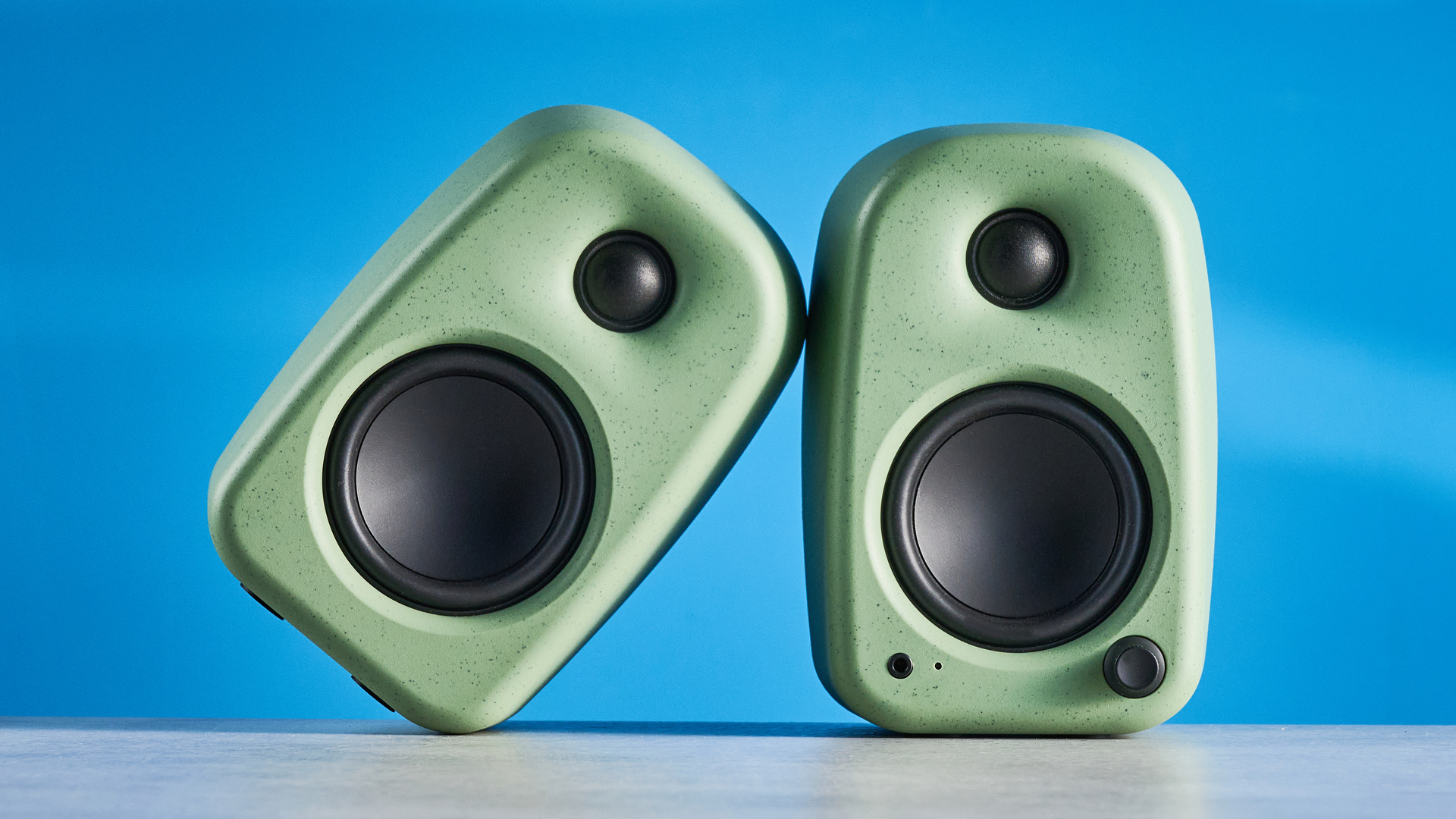 Kanto UKI bookshelf speakers in sage photographed in front of a blue background