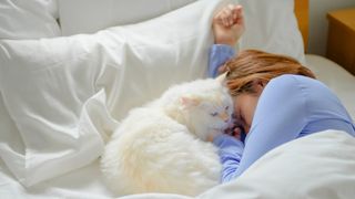 A white cat sits in bed next to a sleeping lady in bed