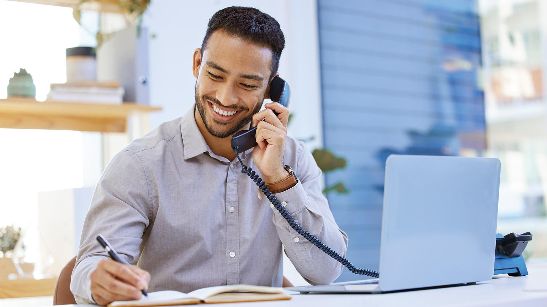A man sitting at a desk writing in a notepad with laptop open and on the phone