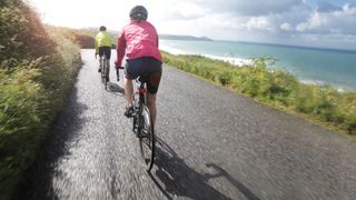 View of woman cycling up a seaside road from behind overlooking the sea