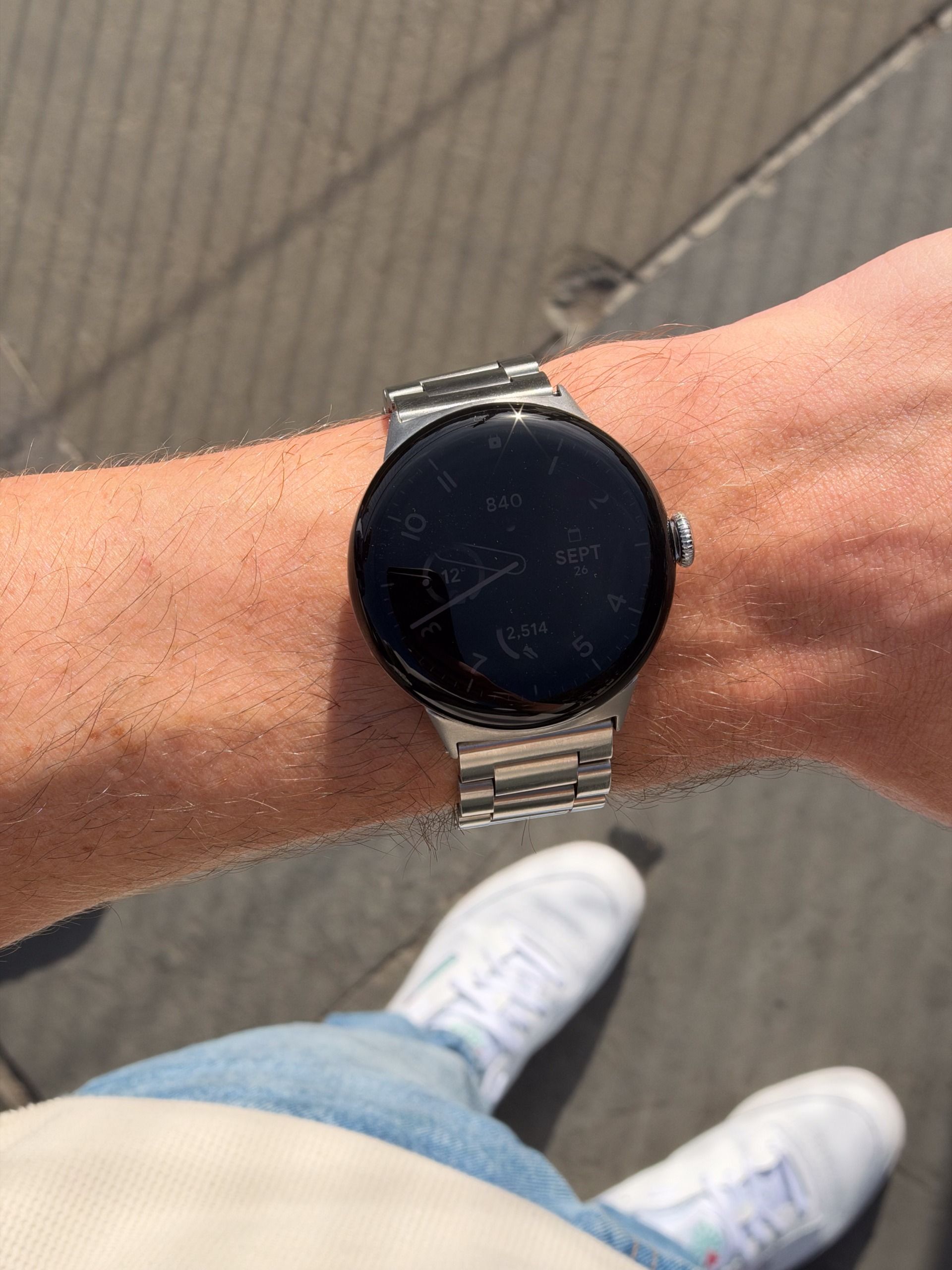 Close-up of a round smartwatch with a dark display and silver metal bracelet on a wrist outdoors.