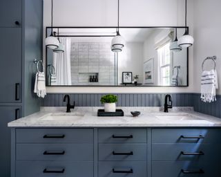A bathroom with blue cabinets, black hardware and pendant lights, and white and grey marble countertop on vanity, with a large mirror behind