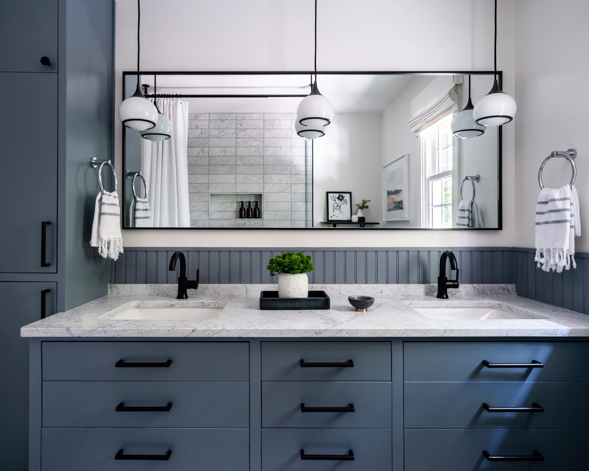 Un baño con gabinetes azules, herrajes negros y luces colgantes, y una encimera de mármol blanco y gris en el tocador, con un gran espejo detrás