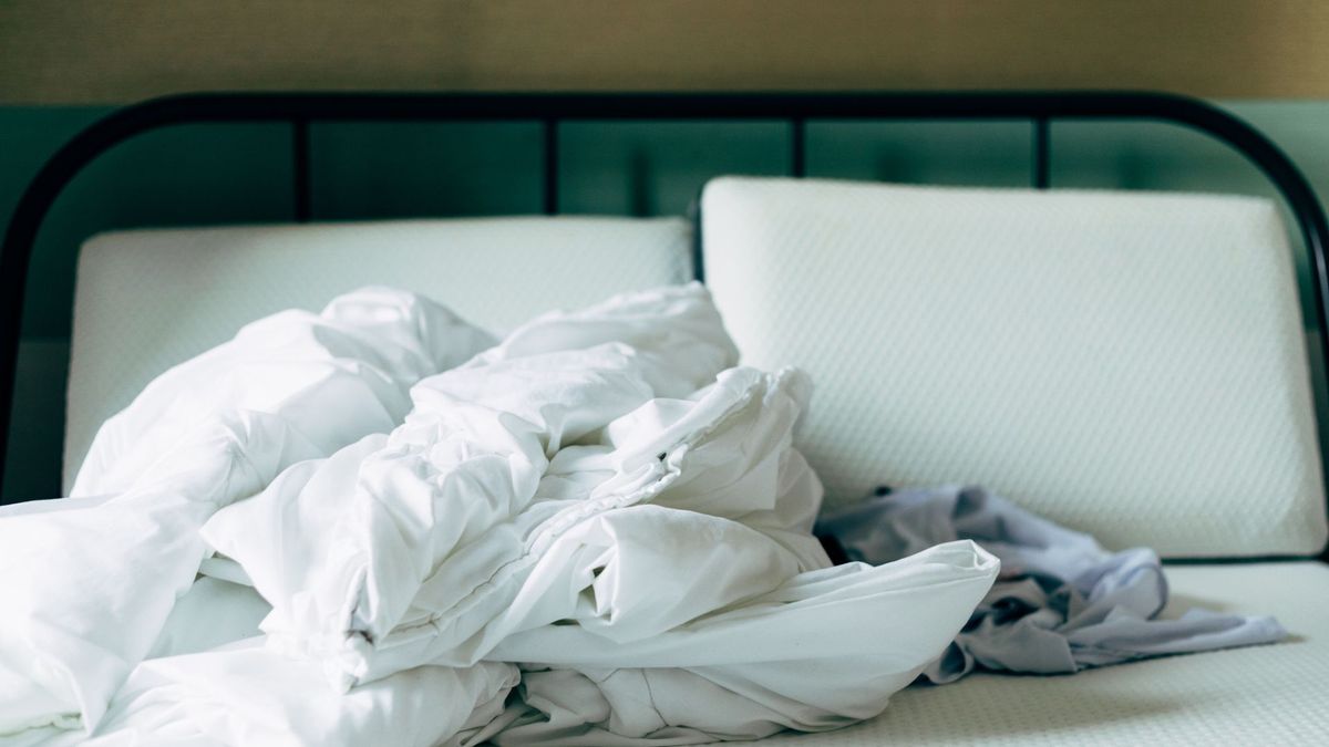 The image shows a bare mattress and pillows and a crumpled white duvet on top of a black bed frame