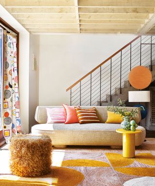 Colorful living room with curvaceous cream sofa, patterned rug, yellow side table, patterned curtains, wooden beams, staircase in background