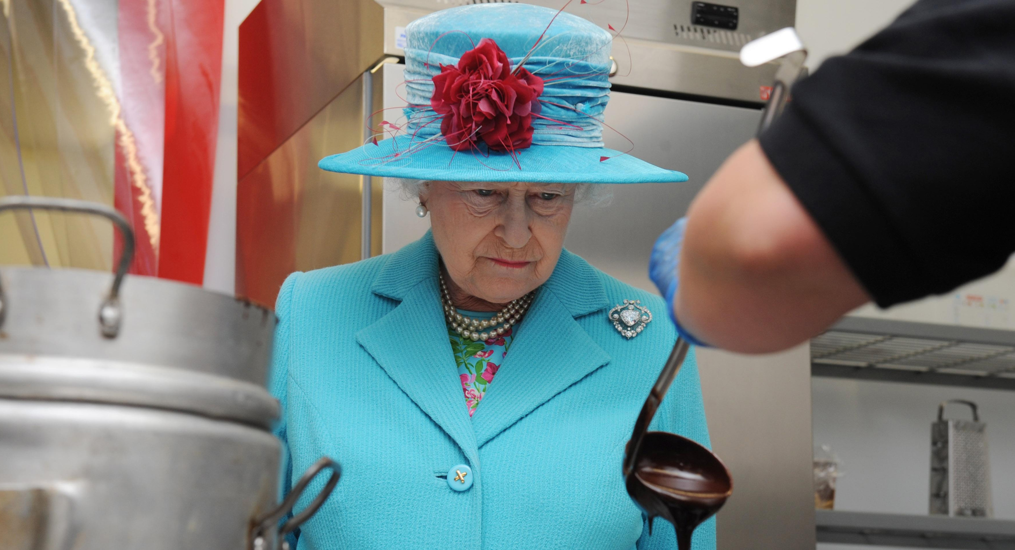 Queen Elizabeth wears a turquoise suit and matching hat and watches a chef serve something with a large metal ladle