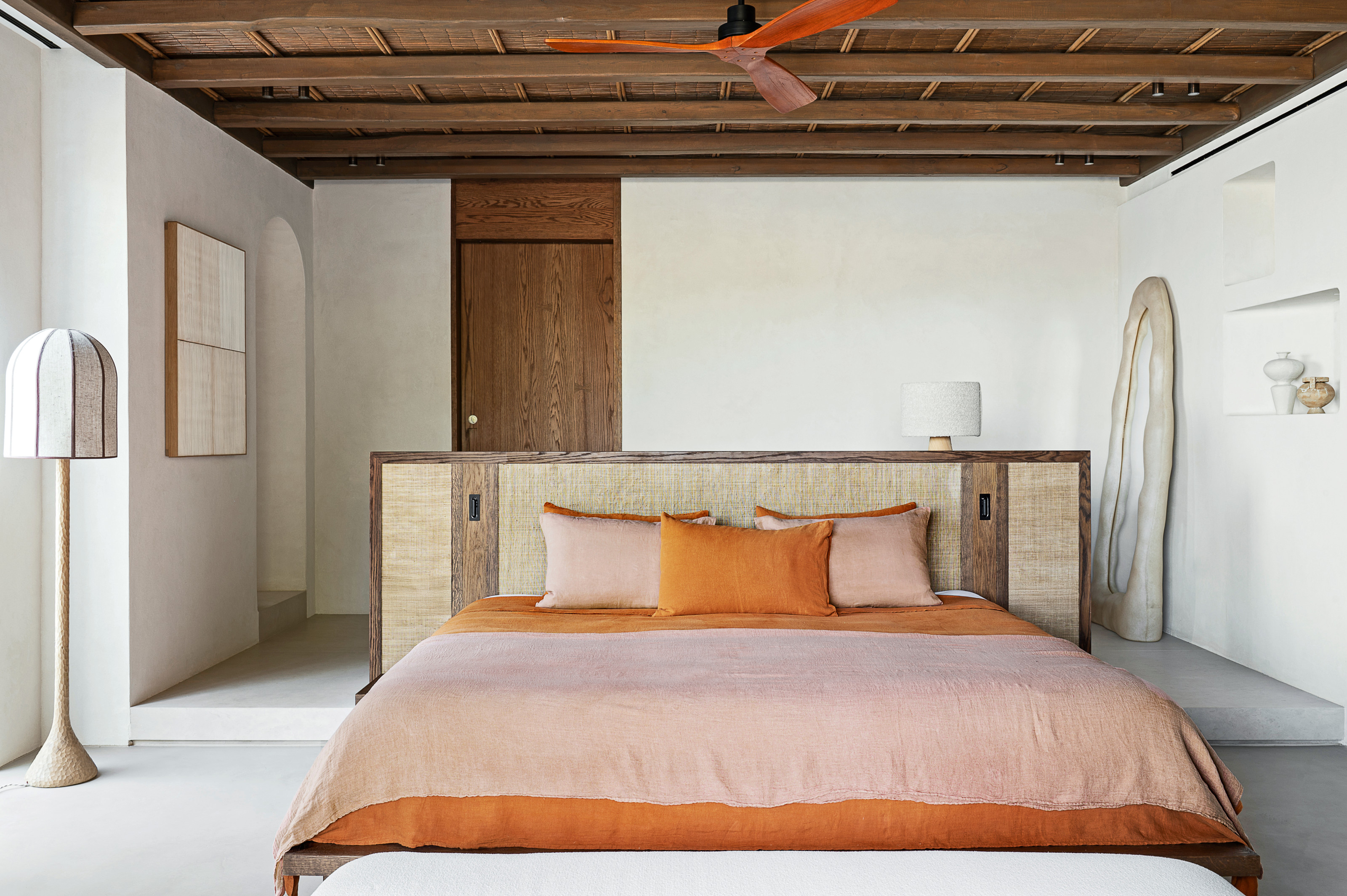 Bed with extended wood and grasscloth headboard in a white room with white table lamp and oversized organic floor mirror
