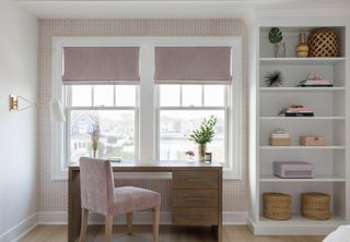 Home office with wooden desk, pink chair and white shelves in front of window