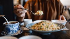 A large bowl of fried rice with vegetables is presented on a dining table while someone lifts a spoonful into their bowl
