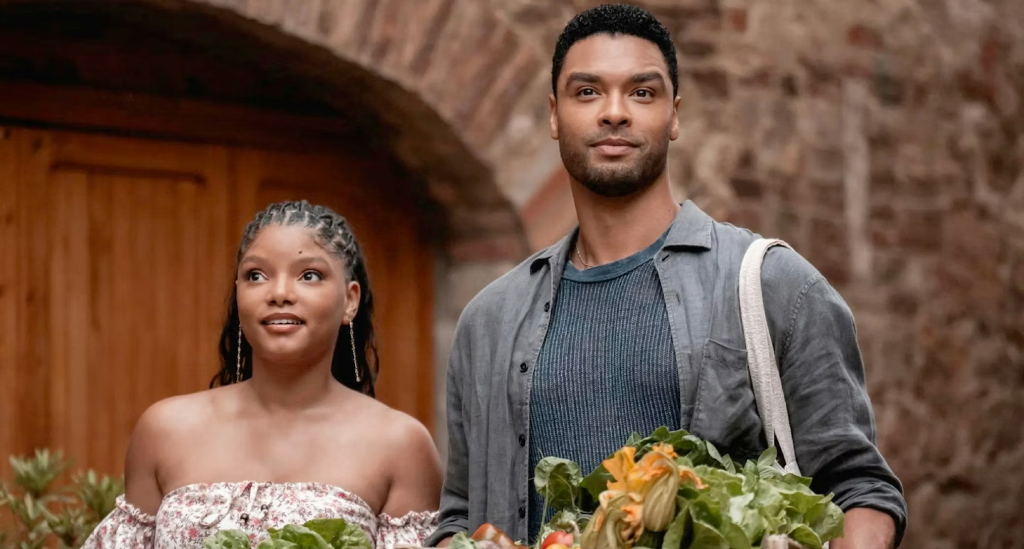 halle bailey and rege jean page smiling on a balcony with produce in front of them in you me and tuscany