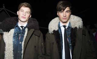 Two male models wearing looks from Dsquared2's collection. Both models are wearing white shirts, black ties, denim jackets and khaki coloured jackets with black and white fur