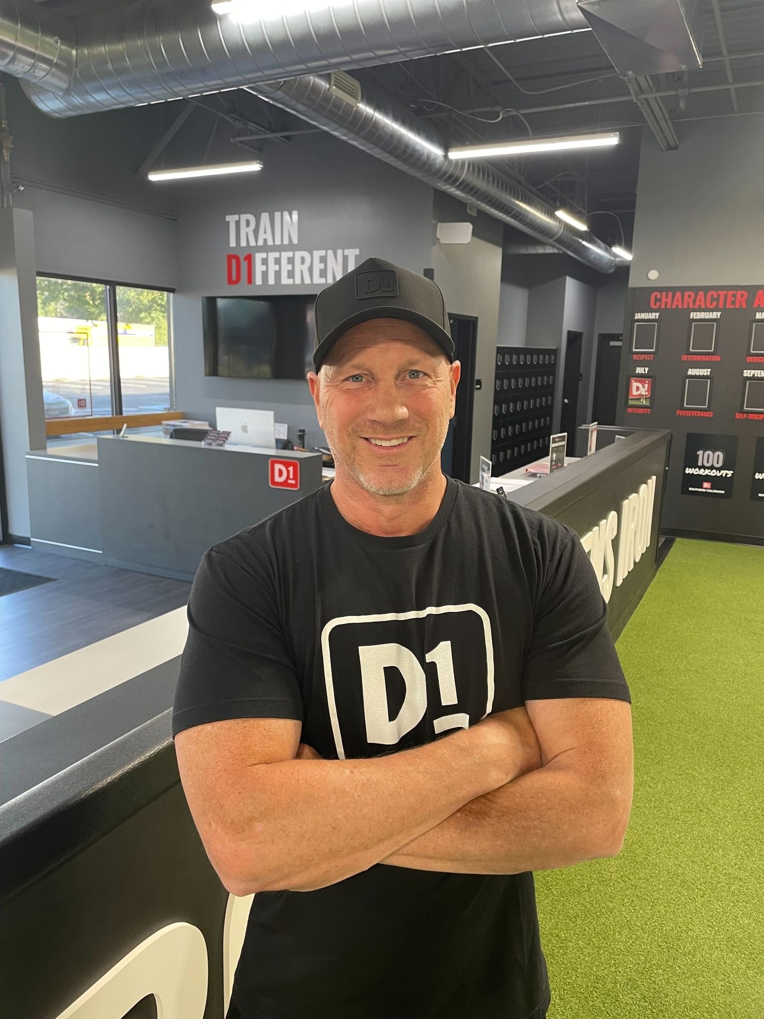 man facing the camera wearing a black tshirt with D1 on it and a black cap. he's folding his arms, smiling and standing in gym entrance.