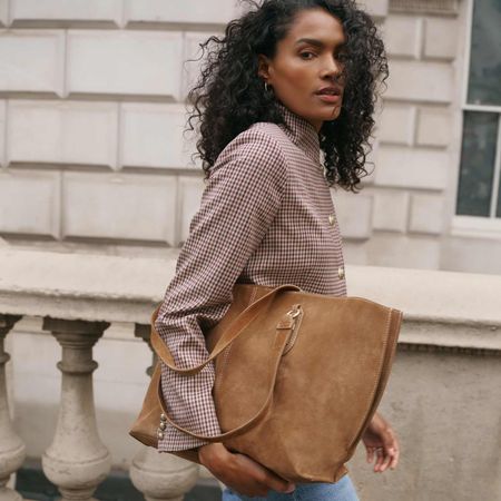 Model wearing a plaid jacket with gold buttons and a camel brown suede tote bag sold at Mint Velvet