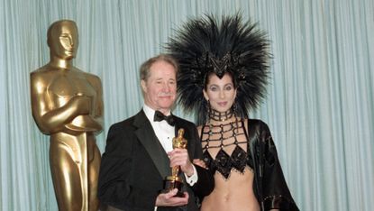 Don Ameche holding the Best Supporting Actor Oscar award for his role in Cocoon, backstage at the Academy Awards with award presenter Cher, who is wearing an unusual Bob Mackie evening gown with a feather headdress.