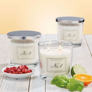A trio of Aldi jar candles arranged on a white wooden surface with a dish of pomegranate seeds, half a lime, half a mandarin and some herb leaves.