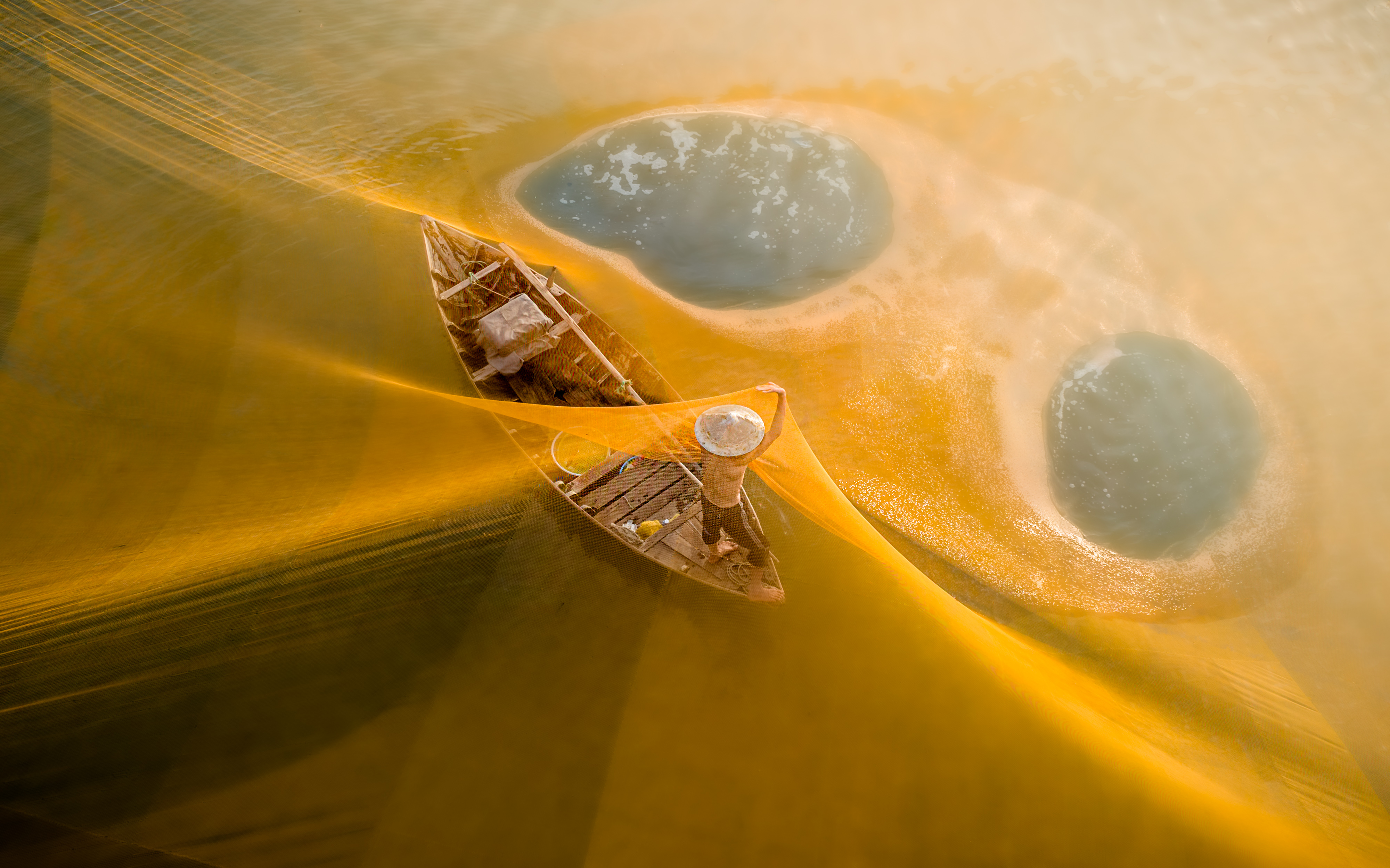 An overhead, high-angle shot captures a fisherman in a conical hat standing on a small wooden boat, surrounded by a massive, glowing golden fishing net stretched across the water