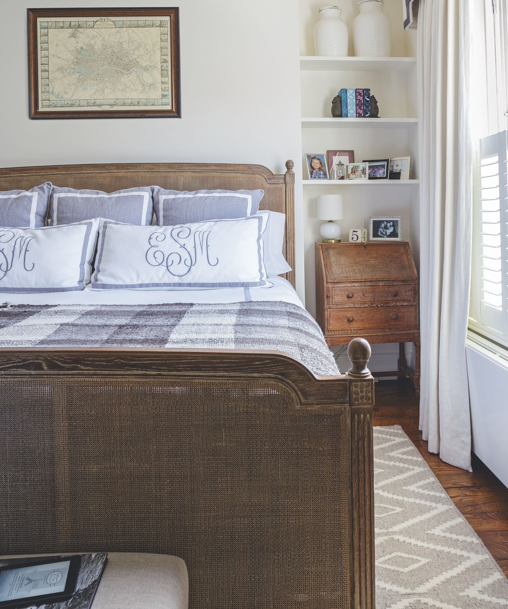 A bright white bedroom with a large rattan bed with grey and white plaid bedding, pillows and cushions seen to the left. Above is a framed map, and at the foot of the bed an upholstered bench is seen with a stack of magazines. Yo the right is a patterned rug seen underneath the bed, a wooden chest of drawers, white shelves with framed pictures, vases and books, and to the right are windows with white blinds and white curtains.