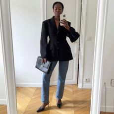 female fashion influencer Sylvie Mus poses for a mirror selfie in a structured, cinch waist black blazer, drop earrings, clutch bag, jeans, and black slingback heels