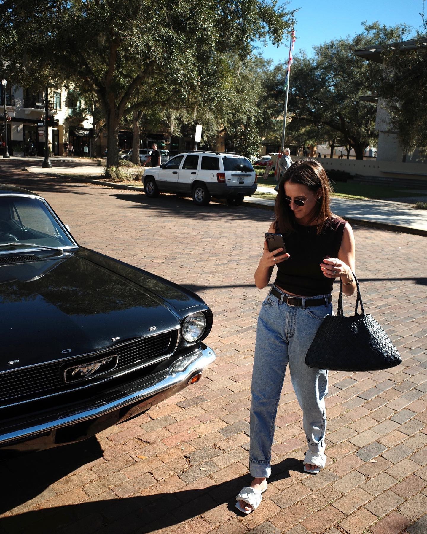 Rachel Whitehouse looking at phone near car wearing black tank top, jeans, belt, and sunglasses