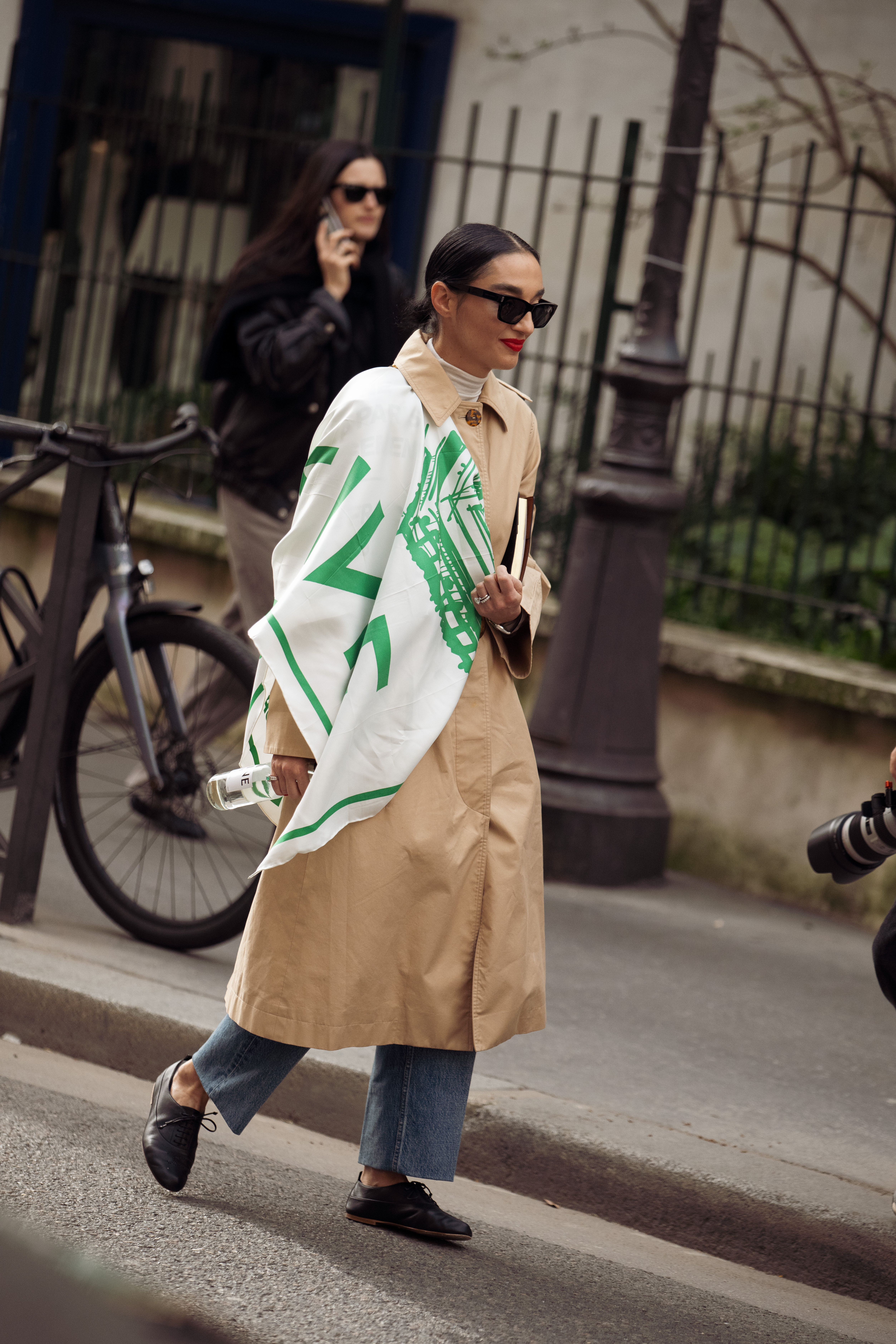 A guest wears a beige trench coat over blue jeans, a white scarf with green graphic, and black leather lace-up derby shoes outside Celine Fall 2026 show during on March 07, 2026 in Paris, France.