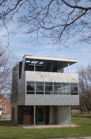 Aluminaire House, front door entrance, glass windows around the top of the building, surrounded by grass and winter time trees, brick building in the distance, blue sky