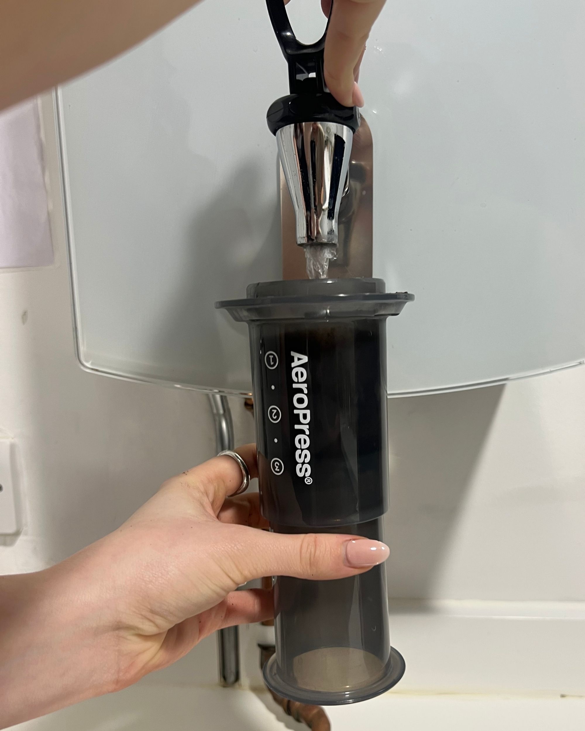 Image of a hand holding a black AeroPress coffee maker under a water spout and hot water going into the maker.