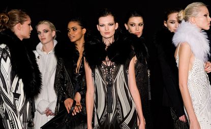 Seven female models wearing black and white pieces from Roberto Cavalli's collection. Some pieces are patterned, some are sleeveless and some feature fur. The background is black