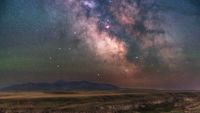 The Milky Way is photographed glowing in a starry night sky above a field with mountains mining the horizon.