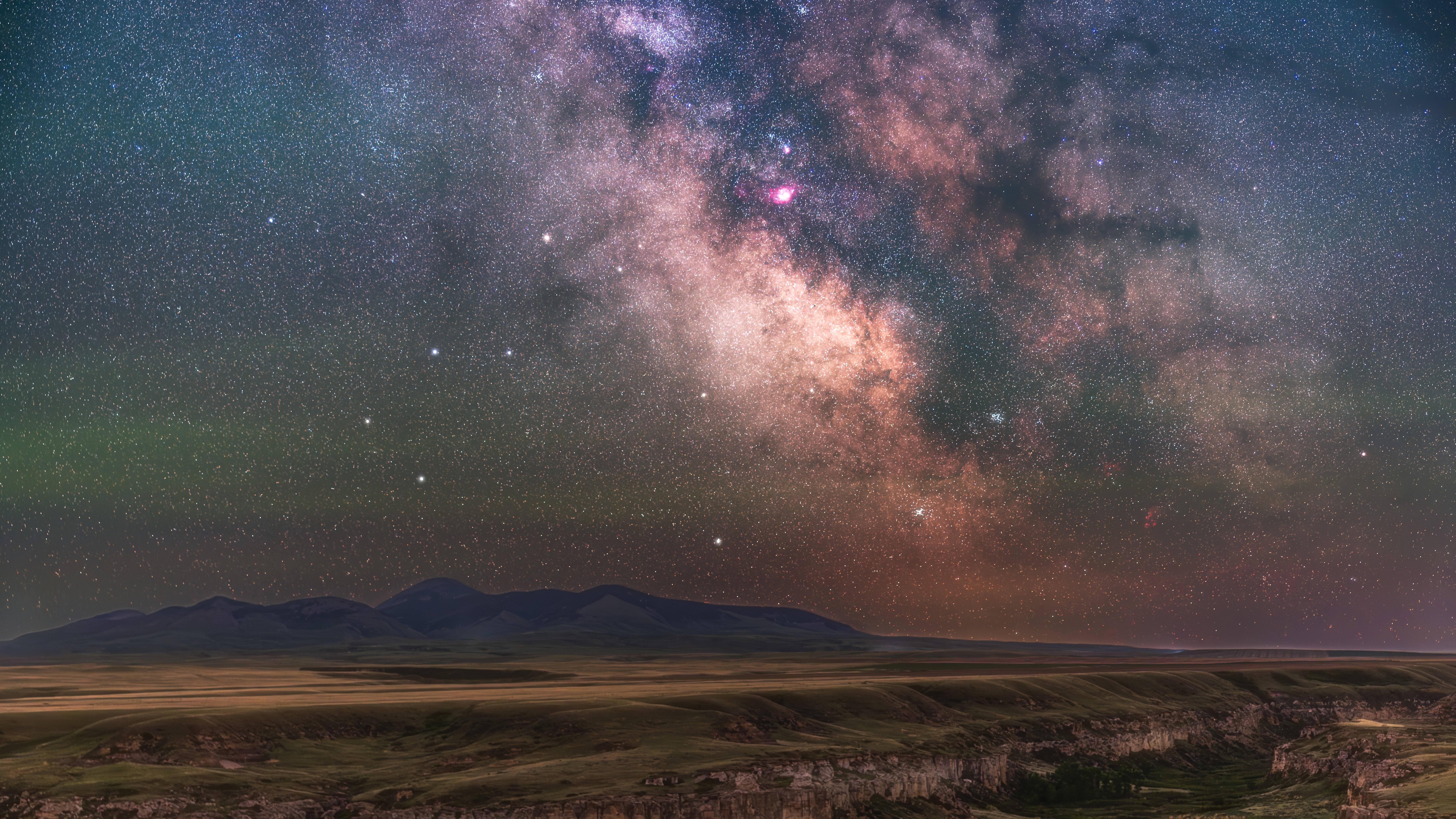 The Milky Way is photographed glowing in a starry night sky above a field with mountains mining the horizon.