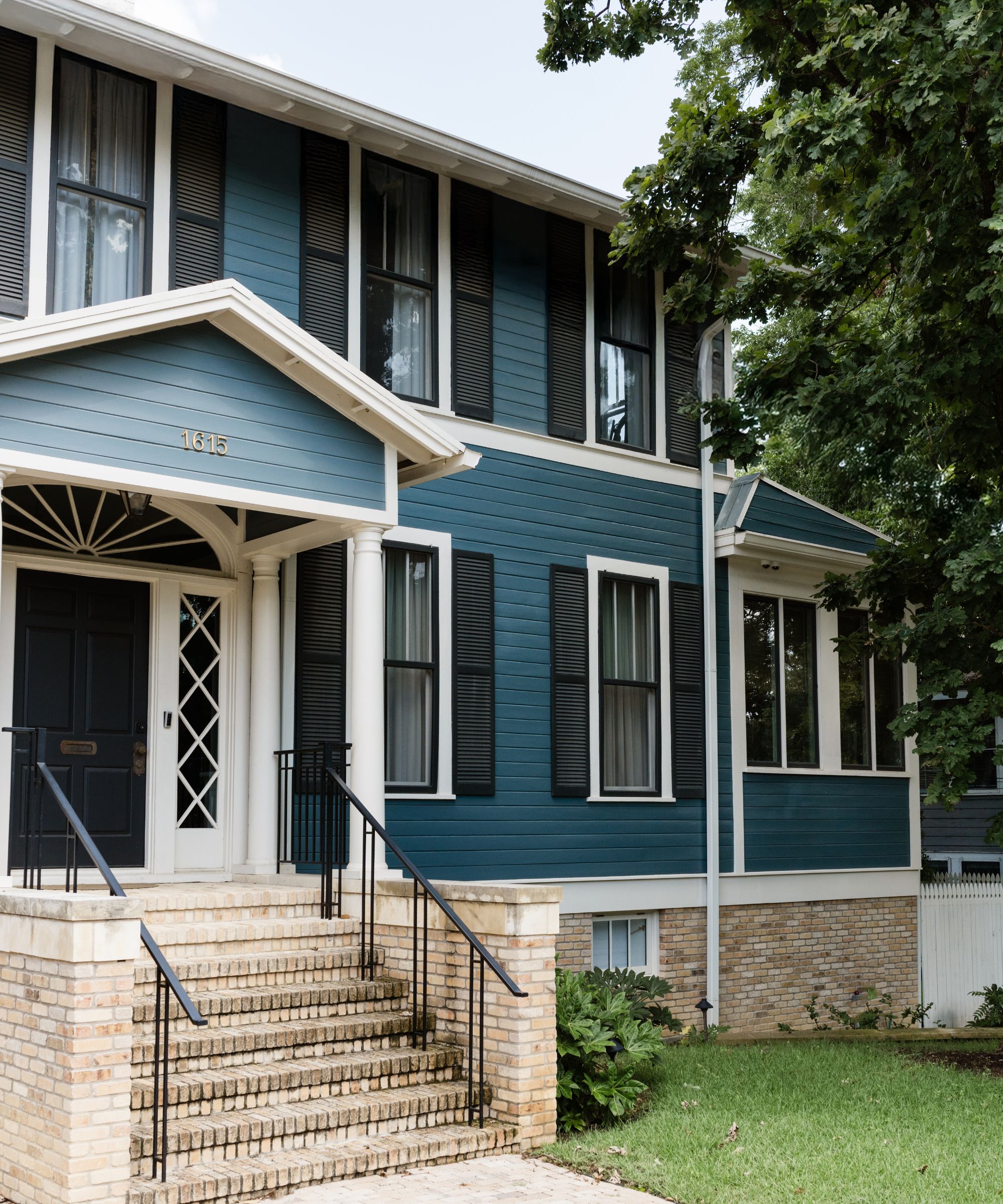 a large historic 1914 home in Austin with blue painted cladding, white windows, black shutters, and a large porch with stone steps