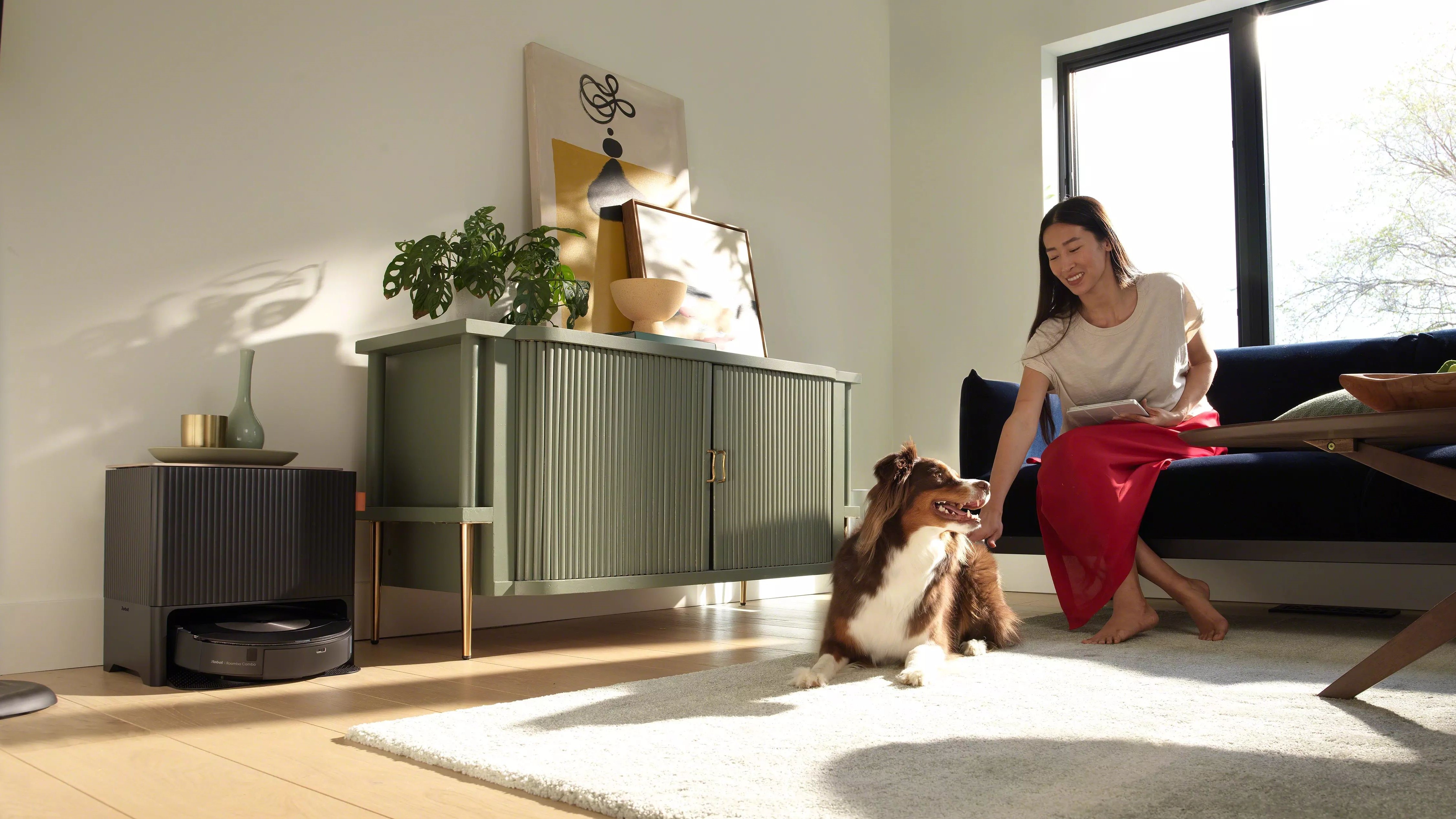 The Roomba Combo j9+ sits in its dock in a person's living room while a person pets their dog on the couch