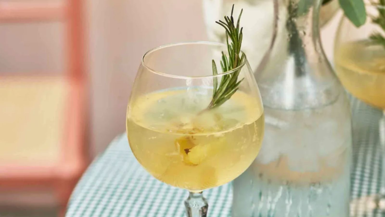 Gin and tonic with lemon and rosemary in a glass with a jug of ice water in the background on a green and white gingham tablecloth