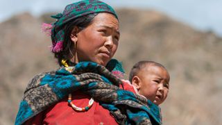 Close-up picture of a Tibetan woman looking into the distance. She is carrying a baby in a shawl on her back. The background of the image is blurry. 