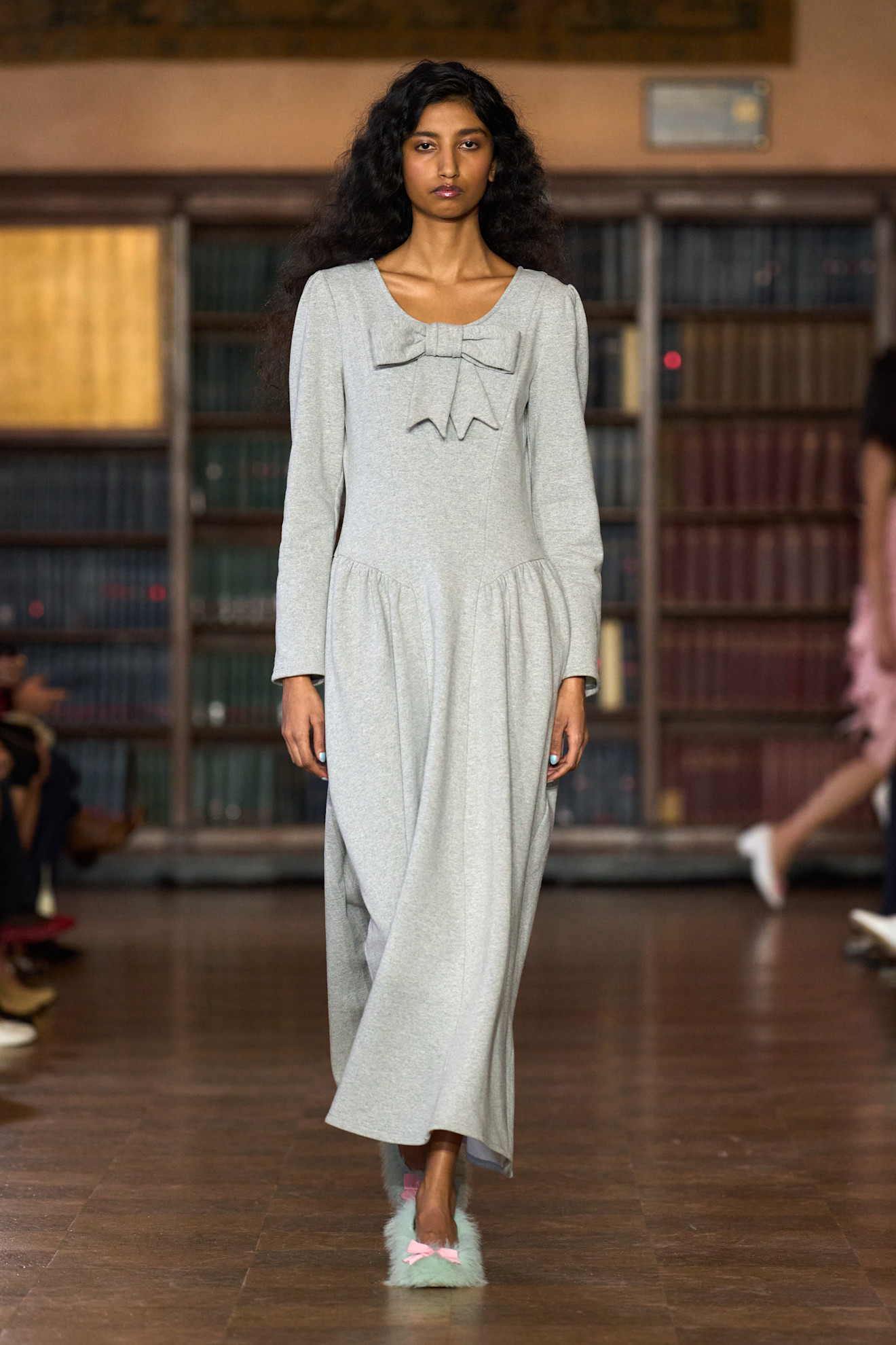 a model on the runway wearing a bow topped dress and furry slipper shoes