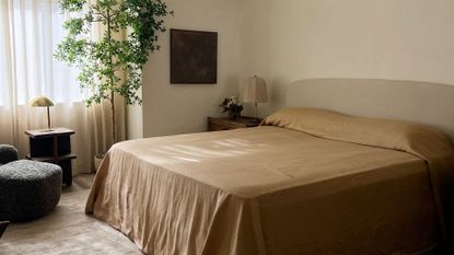 A bedroom with cream walls, a potted tree, a blue footstool, and a bed in sand-colored bedding beside a side table with a table lamp