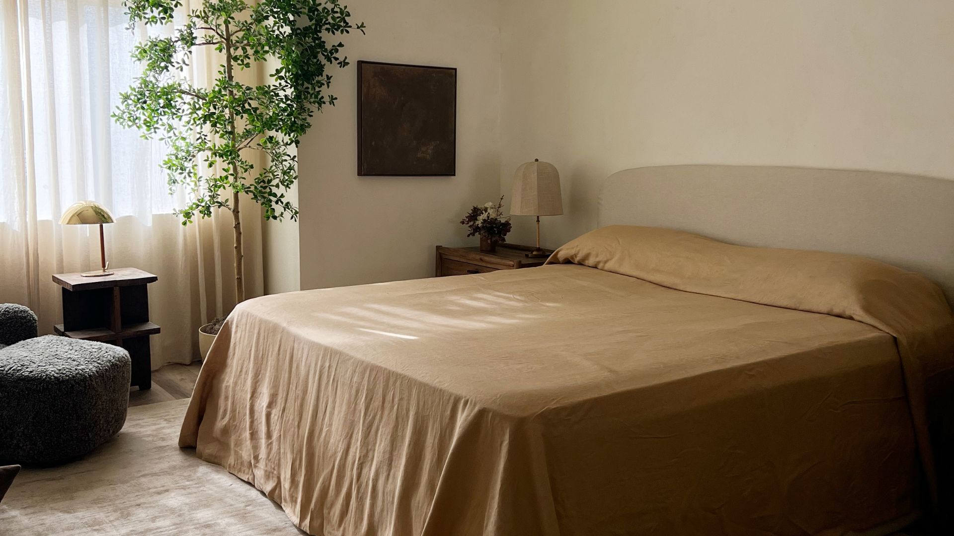 A bedroom with cream walls, a potted tree, a blue footstool, and a bed in sand-colored bedding beside a side table with a table lamp