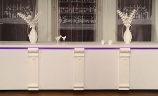 A detailed look at a brasserie-style white 'long bar' constructed from paper stock. On the bar, there are vases with flowers and martini glasses also constructed from paper stock.