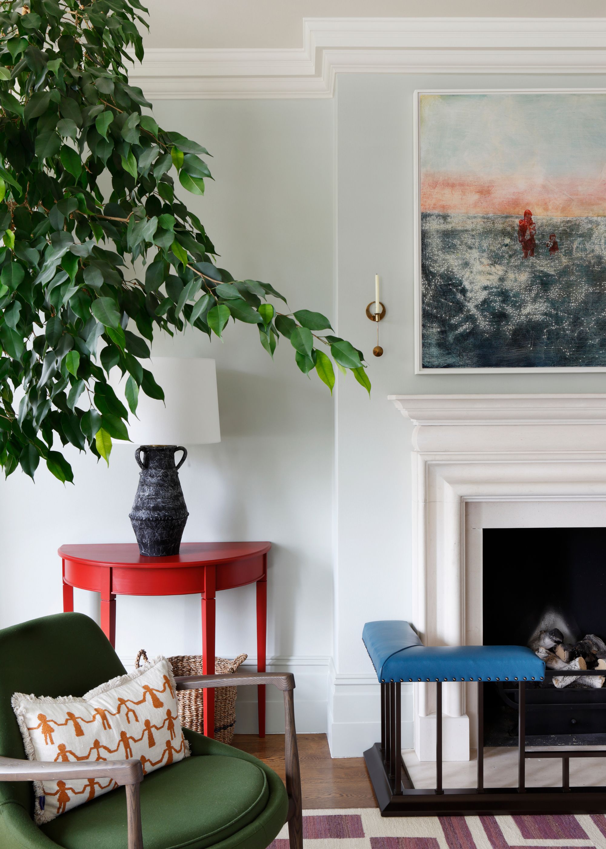 A living room with gray-green walls, a white fireplace with artwork displayed above it, a bright red side table with a lamp, and a moss green accent chair.
