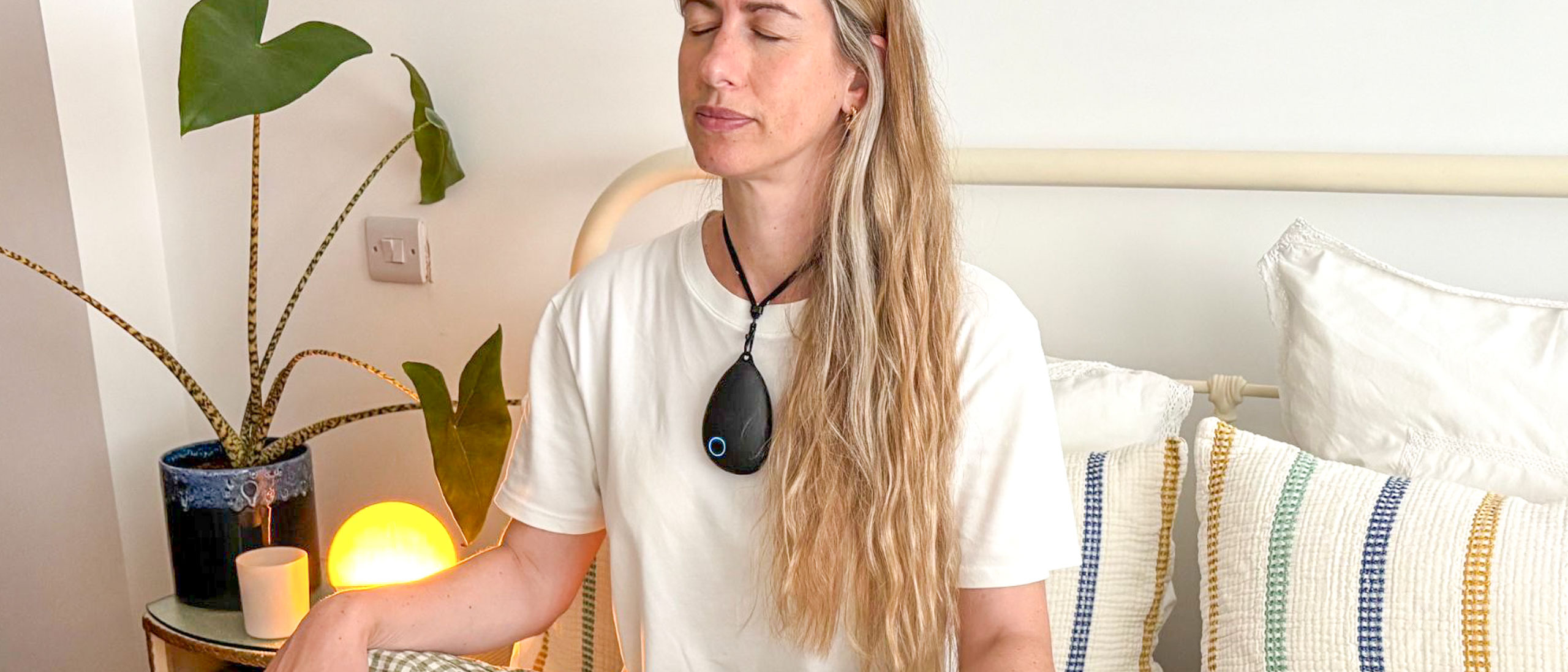 A blond woman with long hair wearing a white t-shirt sits up in bed with her eyes closed and her hands resting on her knees. Around her neck is a black, pebble shaped gadget.